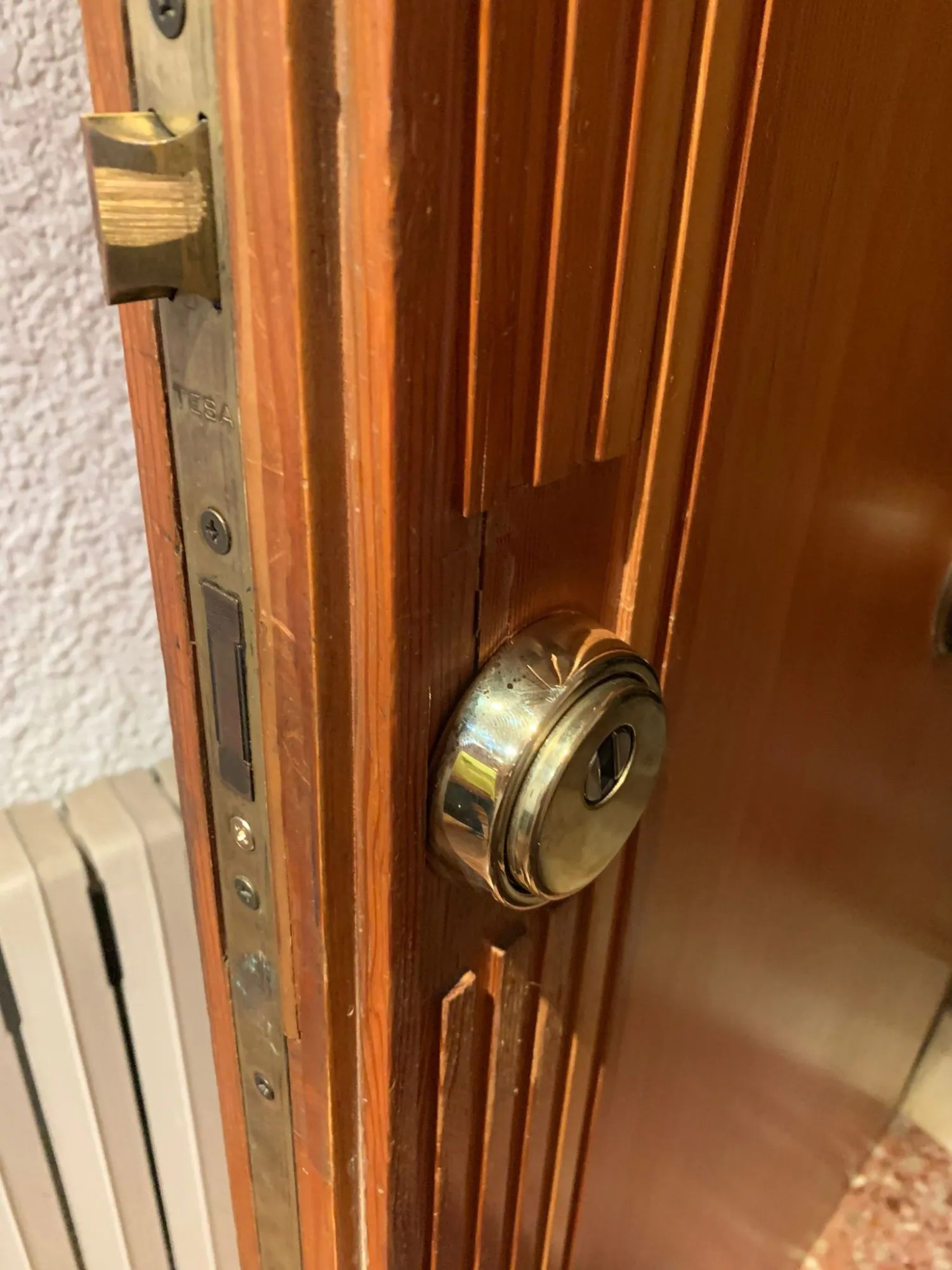Cerradura de embutir TESA en el canto de una puerta de madera en Leganés, con el escudo protector dorado del bombín y la moldura dañada a su lado.