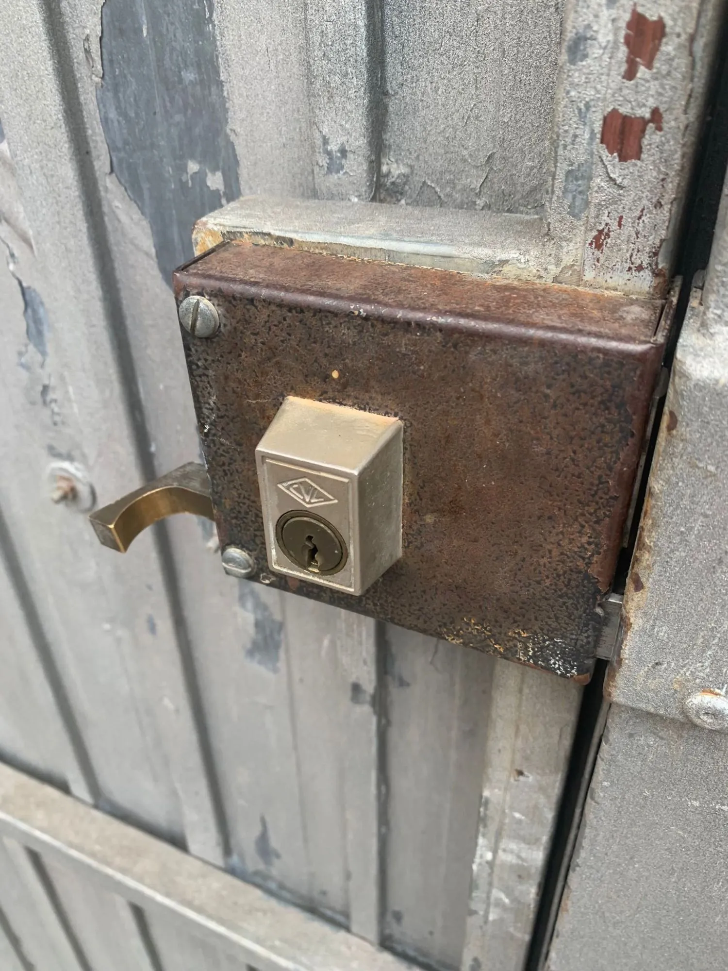 Cerradura de sobreponer CVL oxidada en una puerta metálica gris con la pintura desconchada en Getafe, con su manilla de latón.